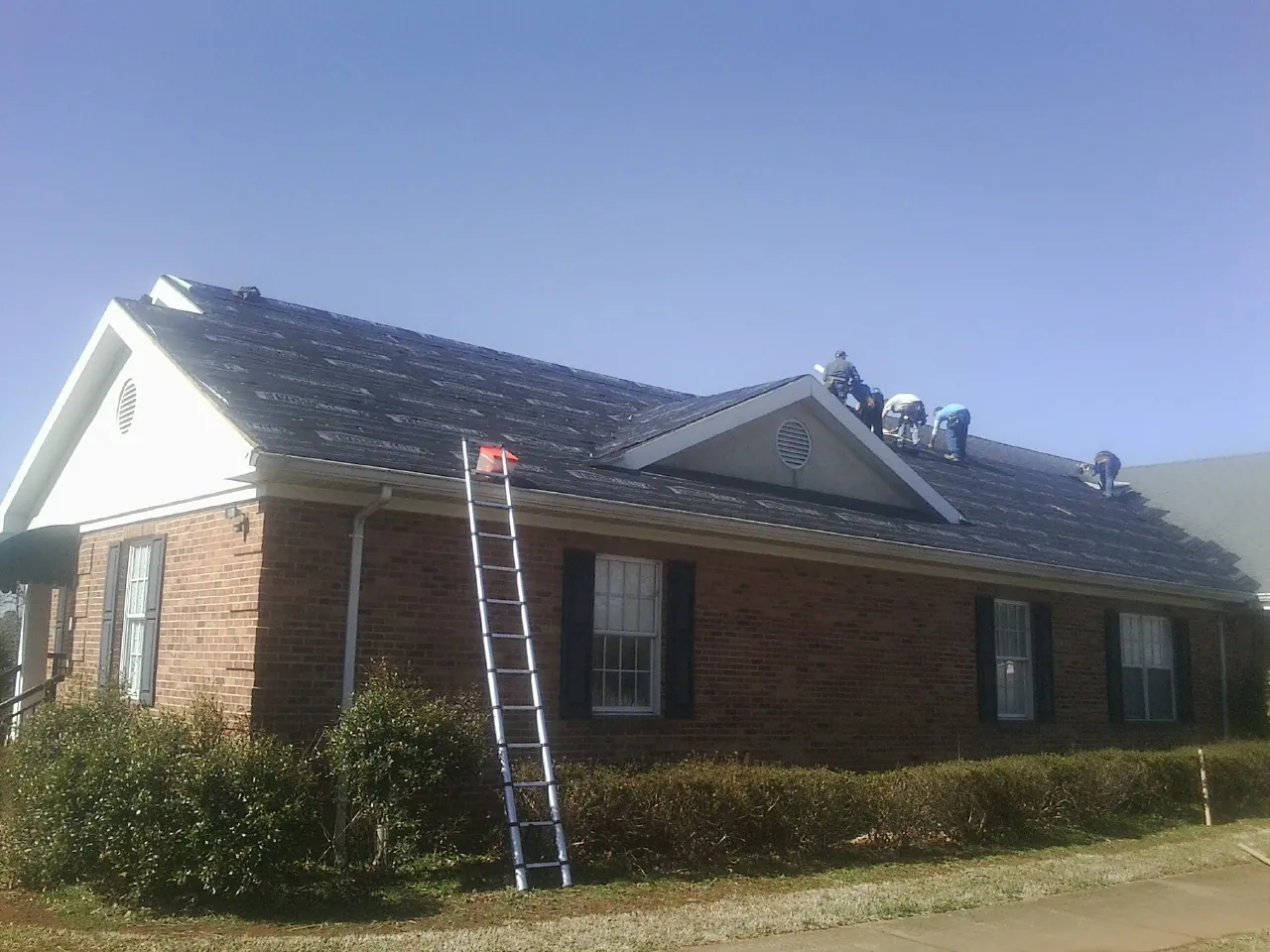Crew tearing shingles off a brick ranch roof with a ladder set — Metal Roof Repair work by our crew in Denham Springs