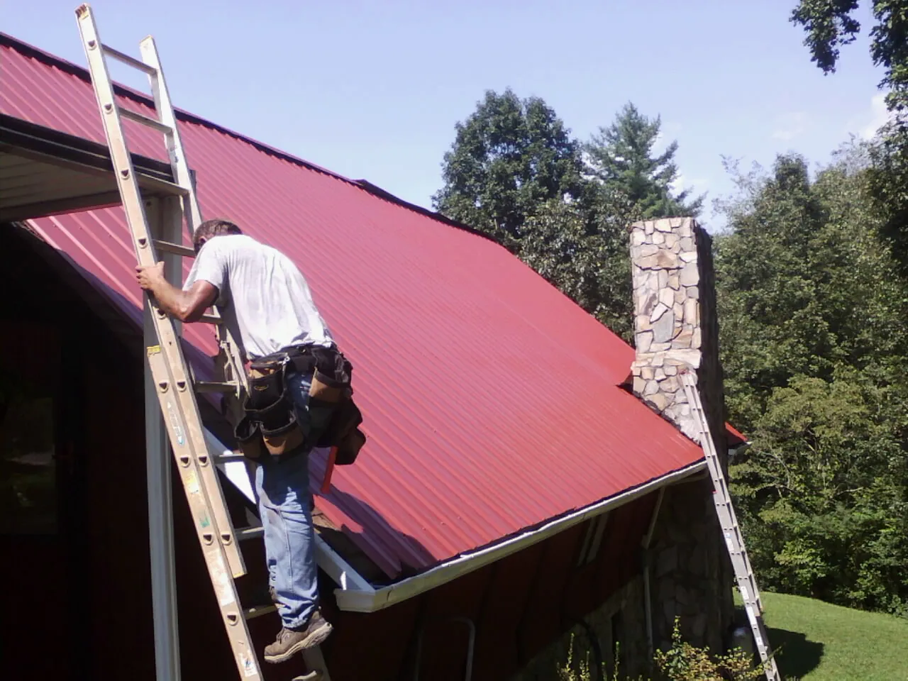 Roofer on a ladder beside a red metal roof and stone chimney — Soffit & Fascia Repair by Roofing Brothers in Denham Springs